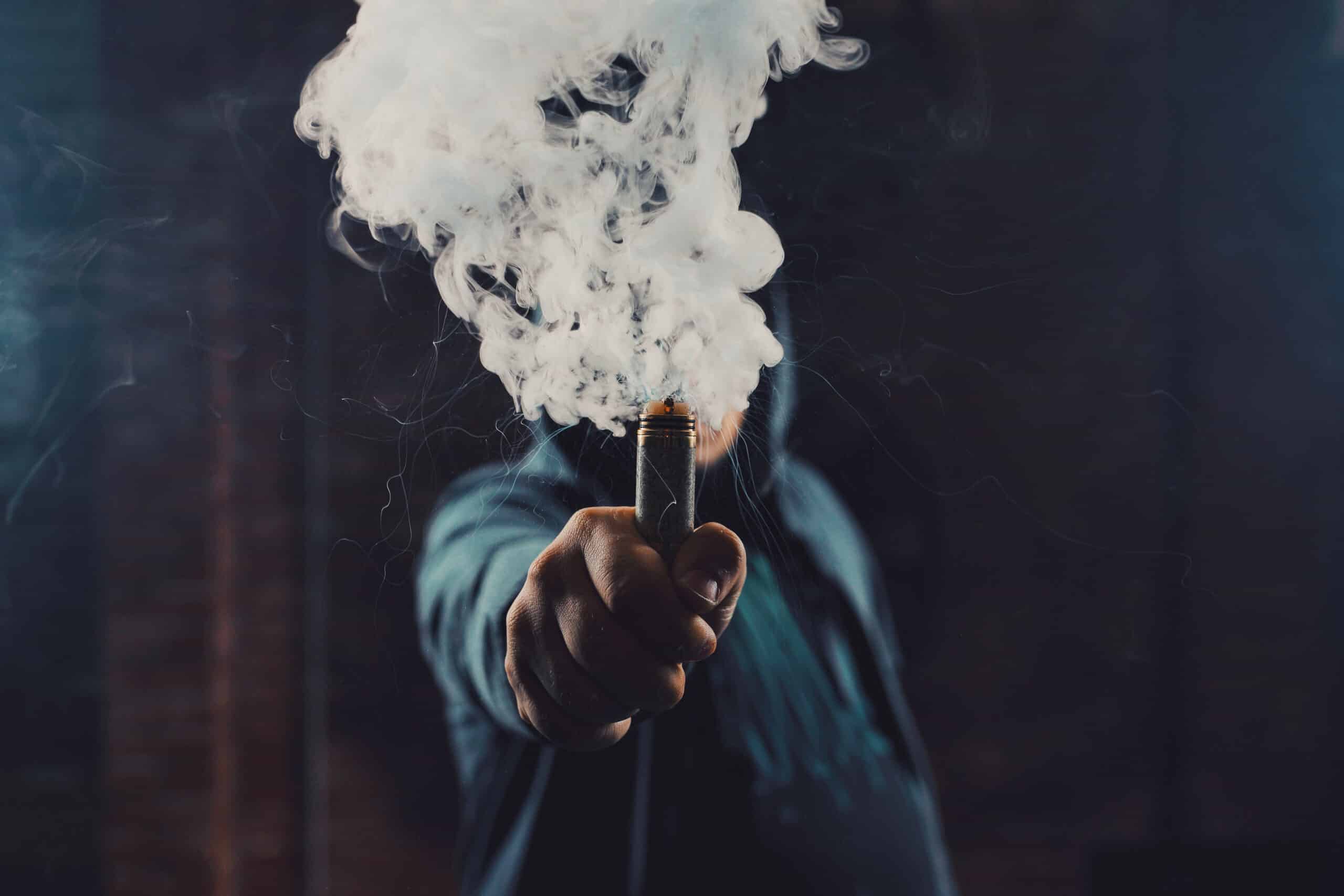 Vaping man wearing a hat, holding up a mod, obscured behind a cloud of vapor.